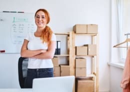 A white, red-head female business owner stands proudly in her office, surrounded by packages